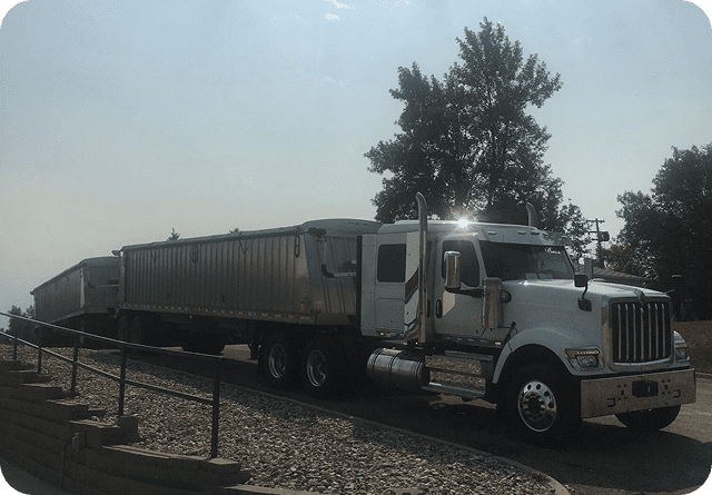 A silver semi-truck parked on a gravel surface under a tree.