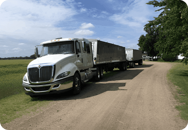 A white semi-truck with a trailer parked on a dirt road near trees.